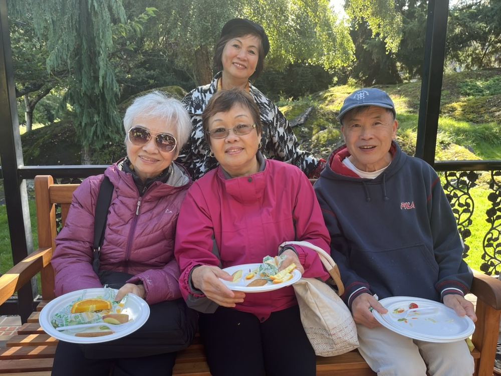 Three Vietnamese seniors sharing food on a bench in a garden, with a VICCIR volunteer behind them